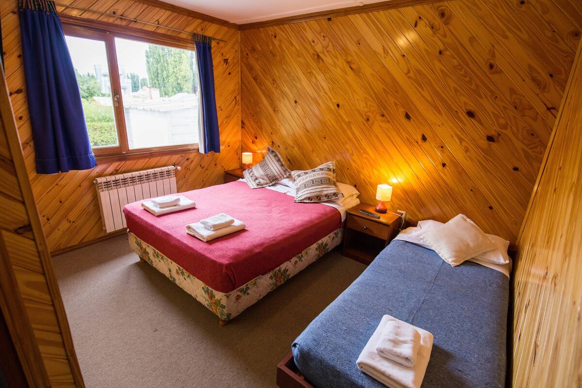 A room with two beds, wooden walls, a window with curtains, and a nightstand with a lamp.