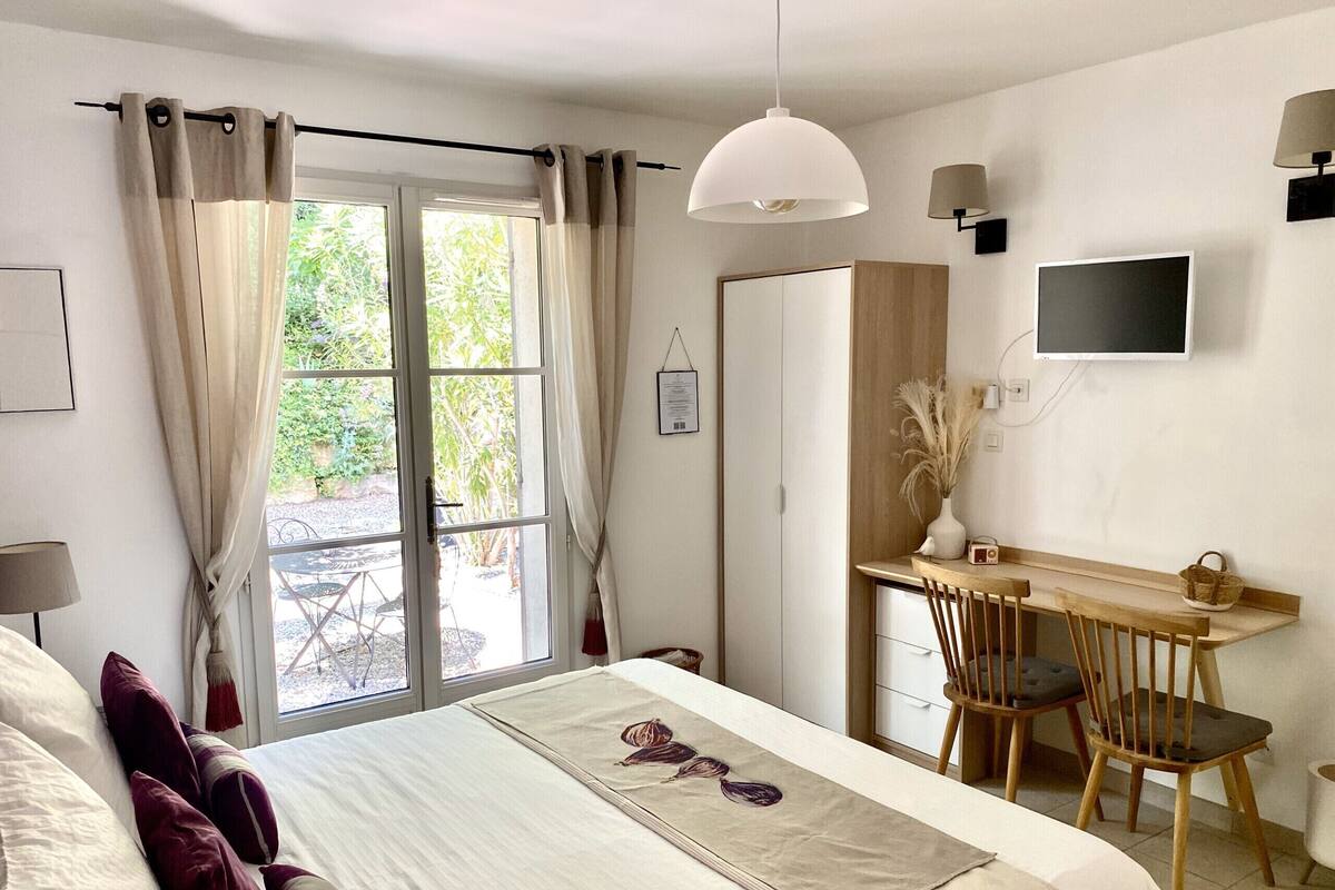 Une chambre avec un lit, un bureau et une chaise. Il y a une fenêtre avec des rideaux, un téléviseur accroché au mur et une lampe.