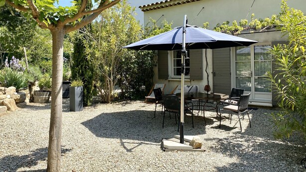 An outdoor patio area with a table and chairs under a large umbrella, surrounded by gravel and various plants.