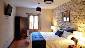 Quarto com cama, mesa de cabeceira, abajur, televisão e parede de pedra.