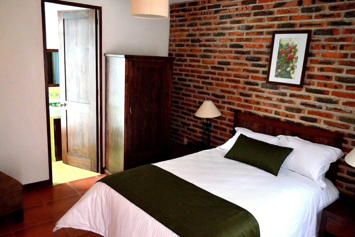 Een slaapkamer met een bakstenen muur, een bed met wit beddengoed, een groene plaid en een houten hoofdbord.