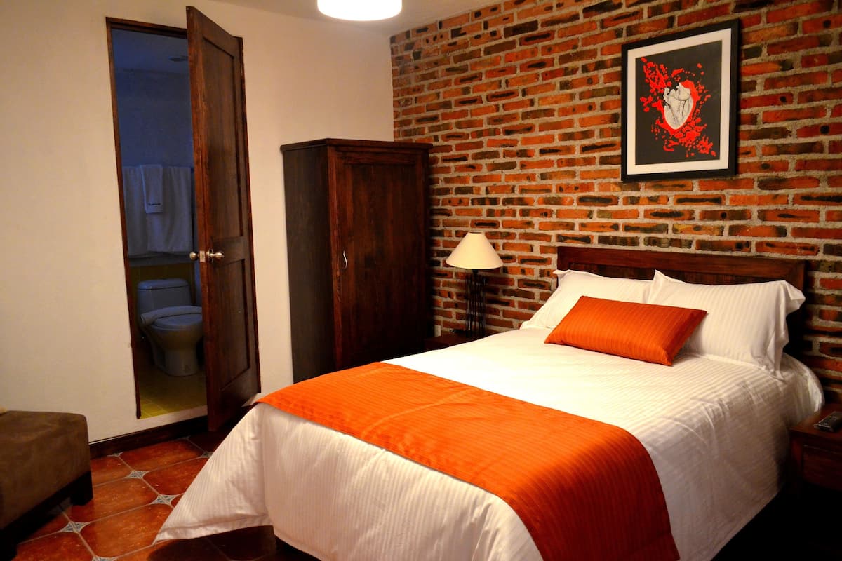 Een slaapkamer met een bakstenen muur, een bed met wit en oranje beddengoed, een houten garderobe, en een zichtbare badkamer door een geopende deur.