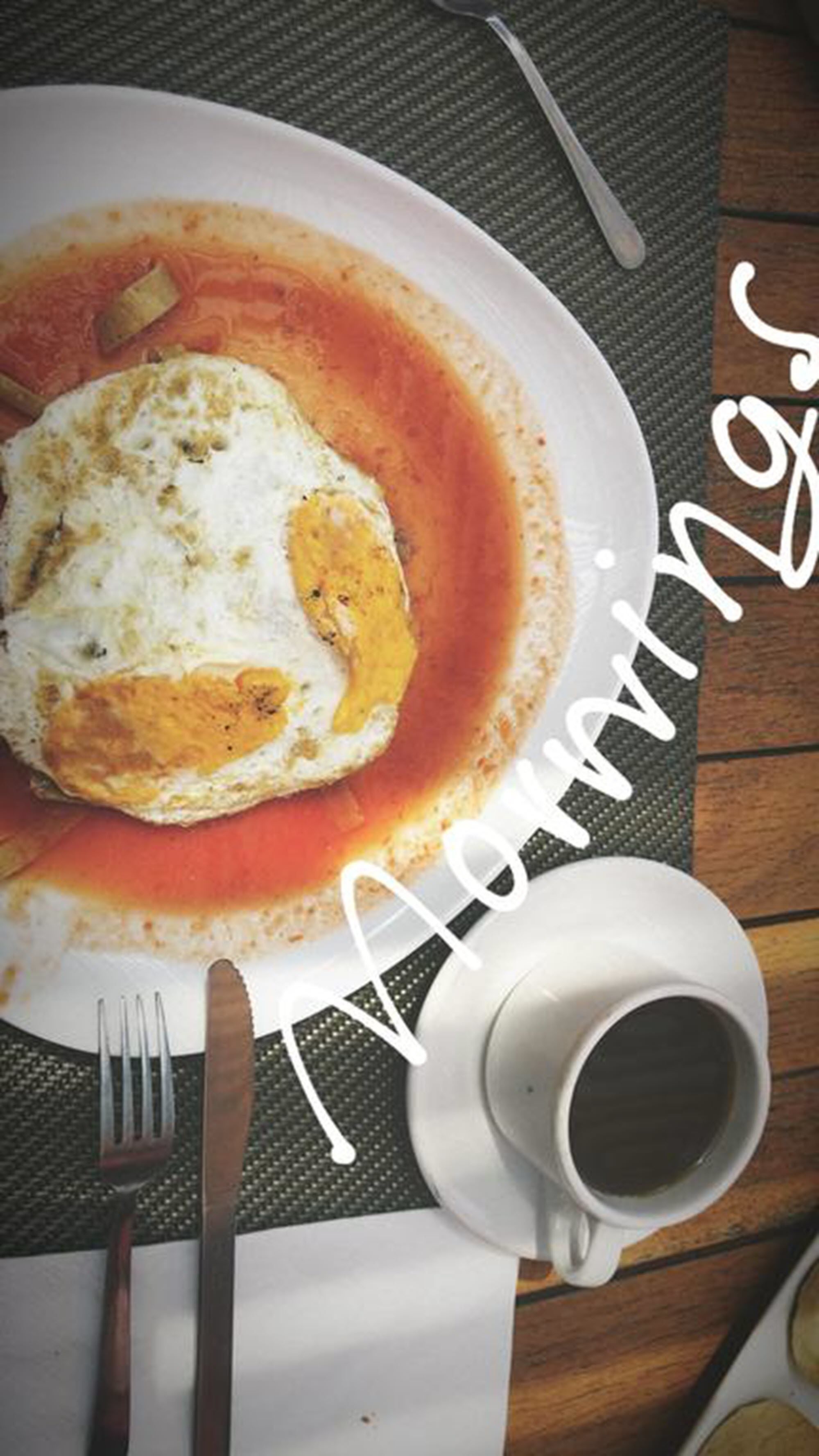 Un plato de desayuno con huevos revueltos y salsa de tomate, una taza de café y cubiertos sobre una mesa con un mantel individual.