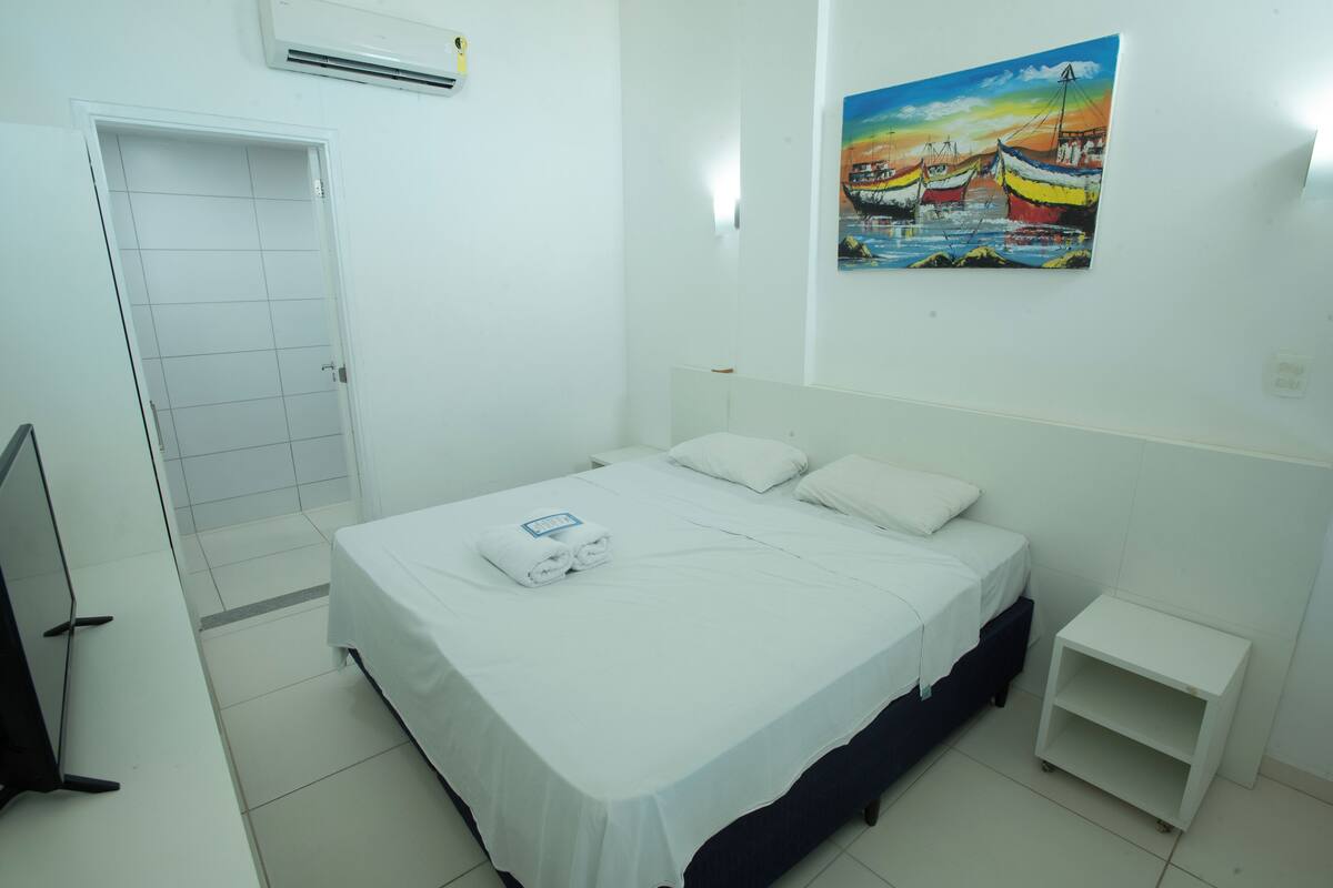 A hotel room with a bed, a small bedside table, a door, and a painting on the wall.