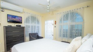 A bedroom with a bed, a dresser, a chair, a television, and a window with curtains. - Columbus Heights Ocean View Large Studio (Ocho Rios)