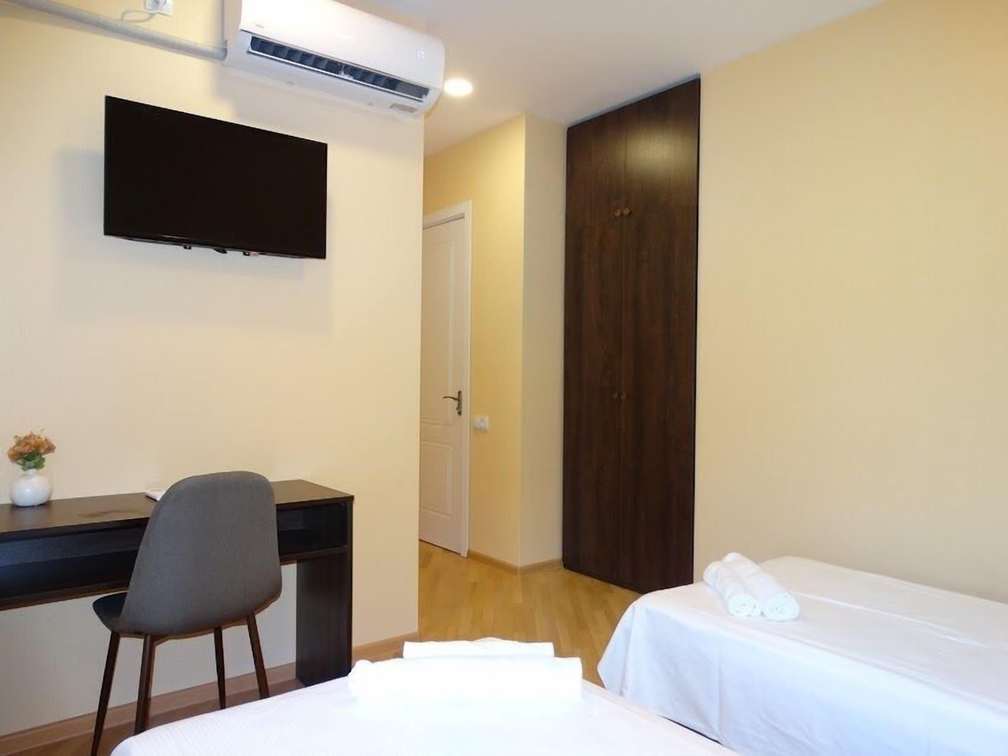 A hotel room with a bed, a desk with a chair, a TV mounted on the wall, and a wardrobe.