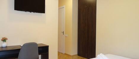 A hotel room with a bed, a desk with a chair, a TV mounted on the wall, and a wardrobe.