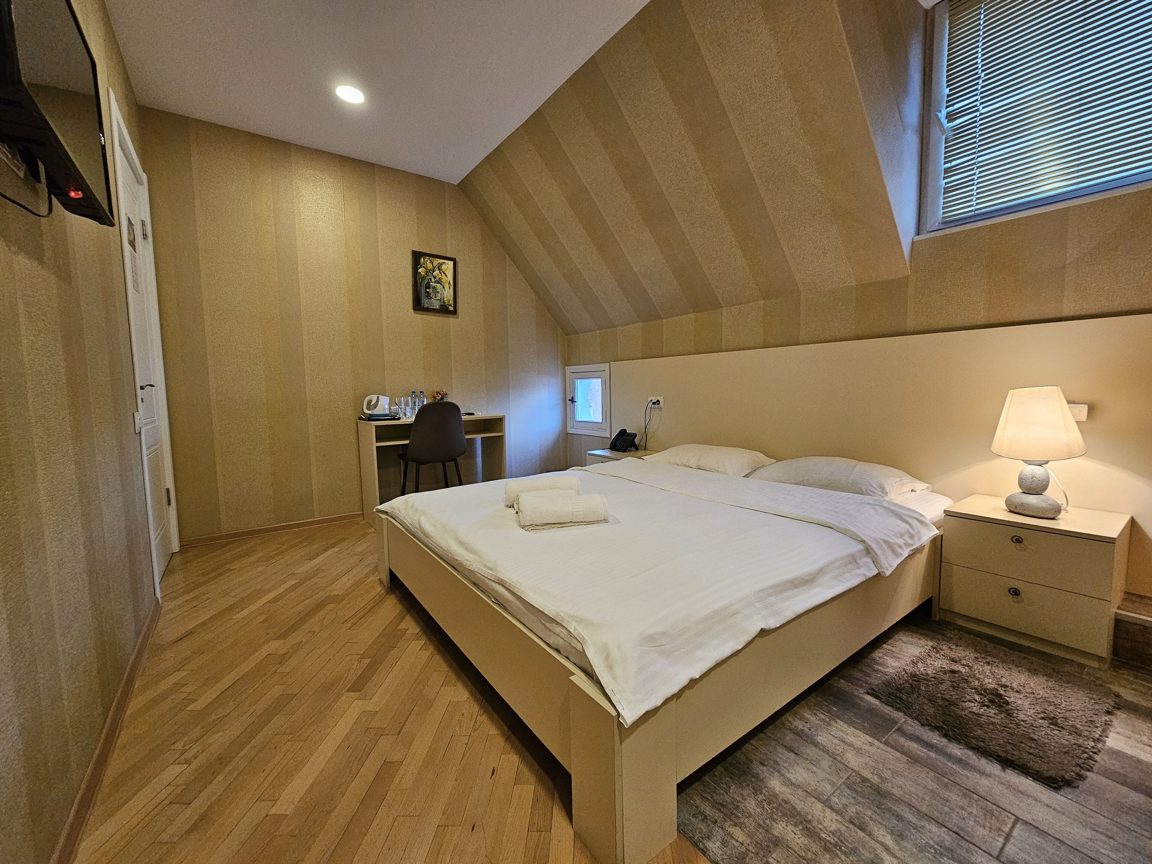 A bedroom with a bed, bedside table, lamp, desk, chair, and a window with blinds.