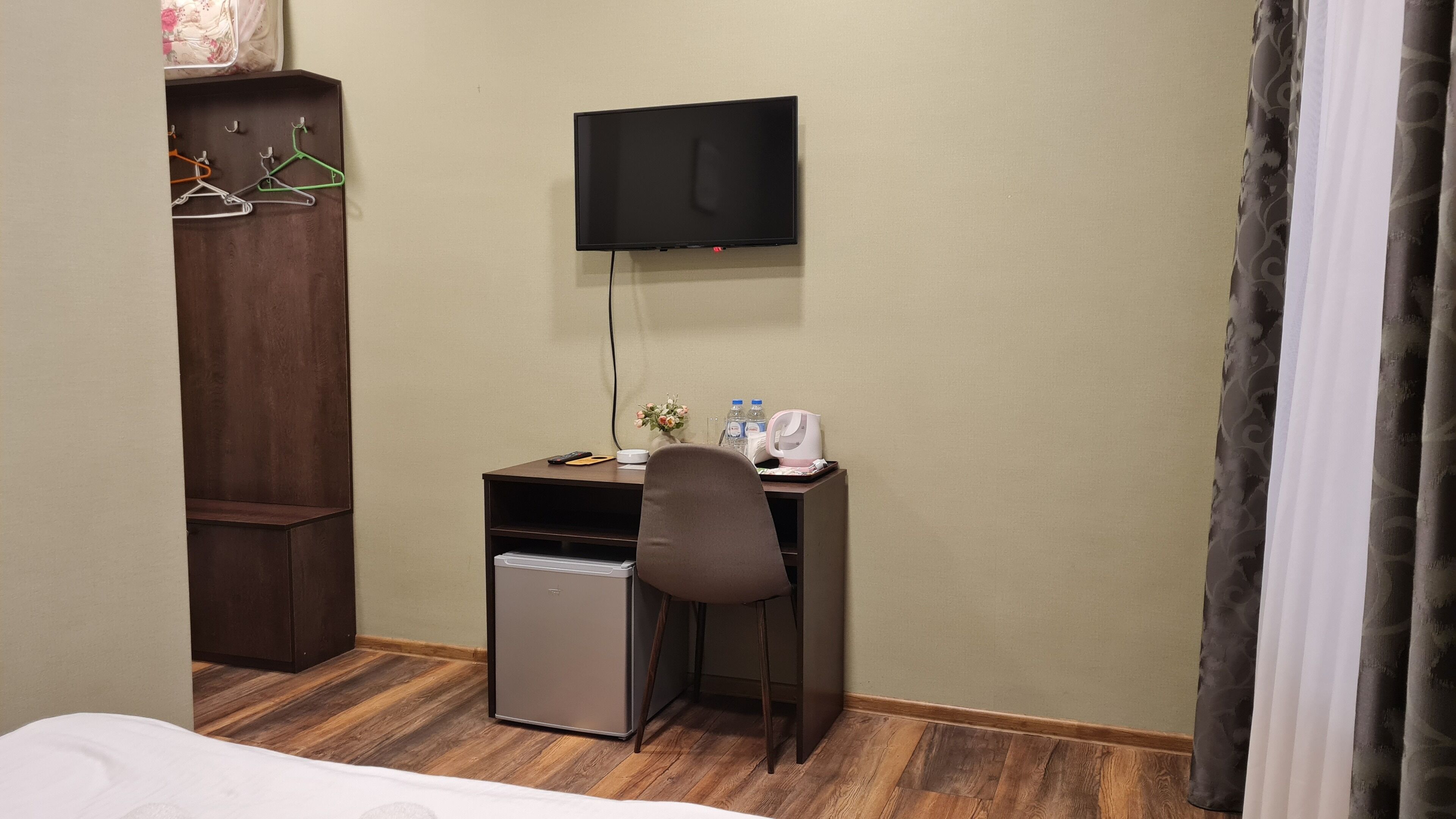 Ein Hotelzimmer mit Bett, Schreibtisch mit Stuhl, Fernseher, kleinem Kühlschrank, Holzschrank und Vorhang.