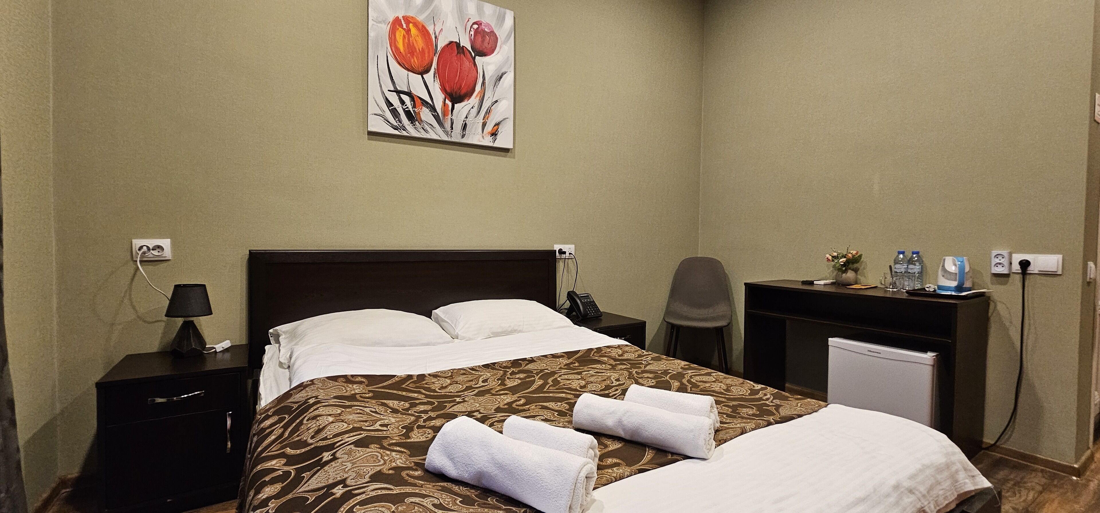A hotel room with a bed, a nightstand, a desk, a chair, a lamp, a painting, and a mini-fridge.
