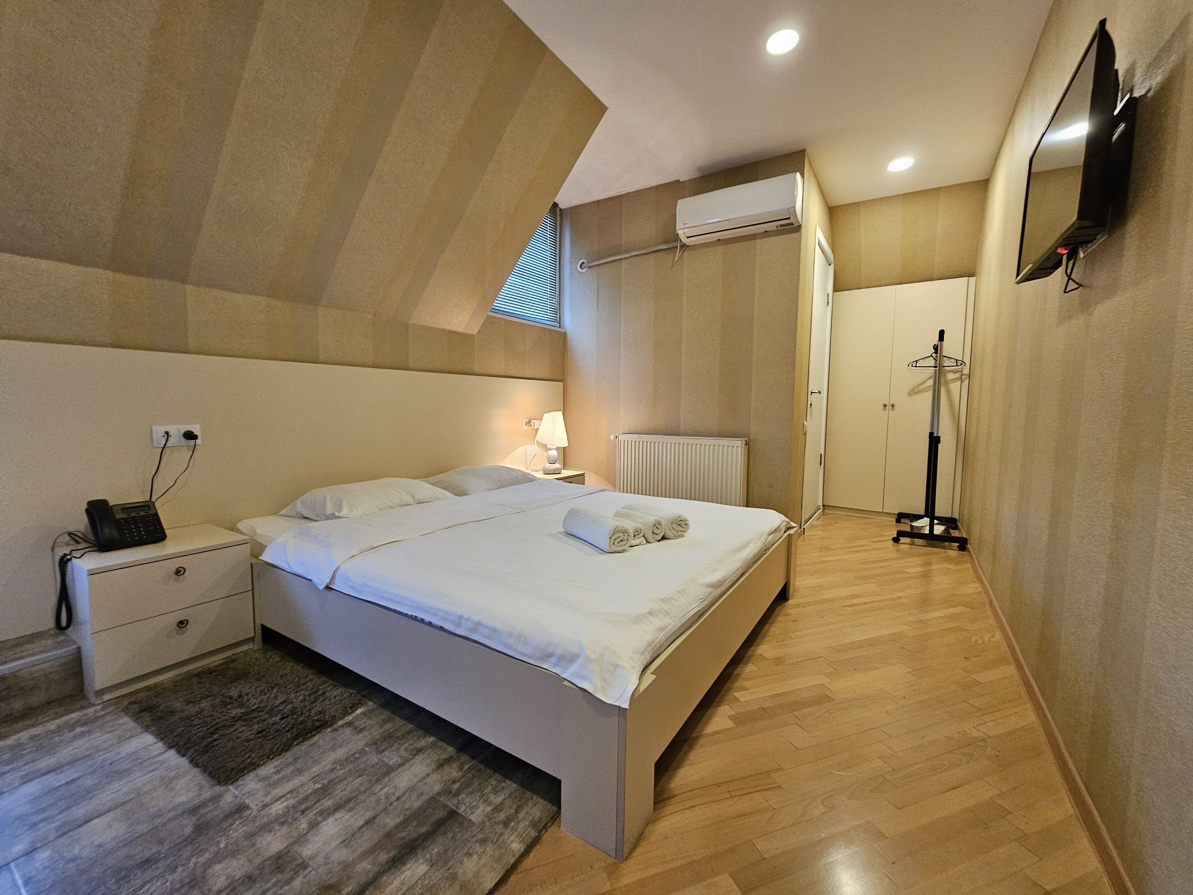 A bedroom with a bed, bedside table, lamp, air conditioning unit, and a coat rack.