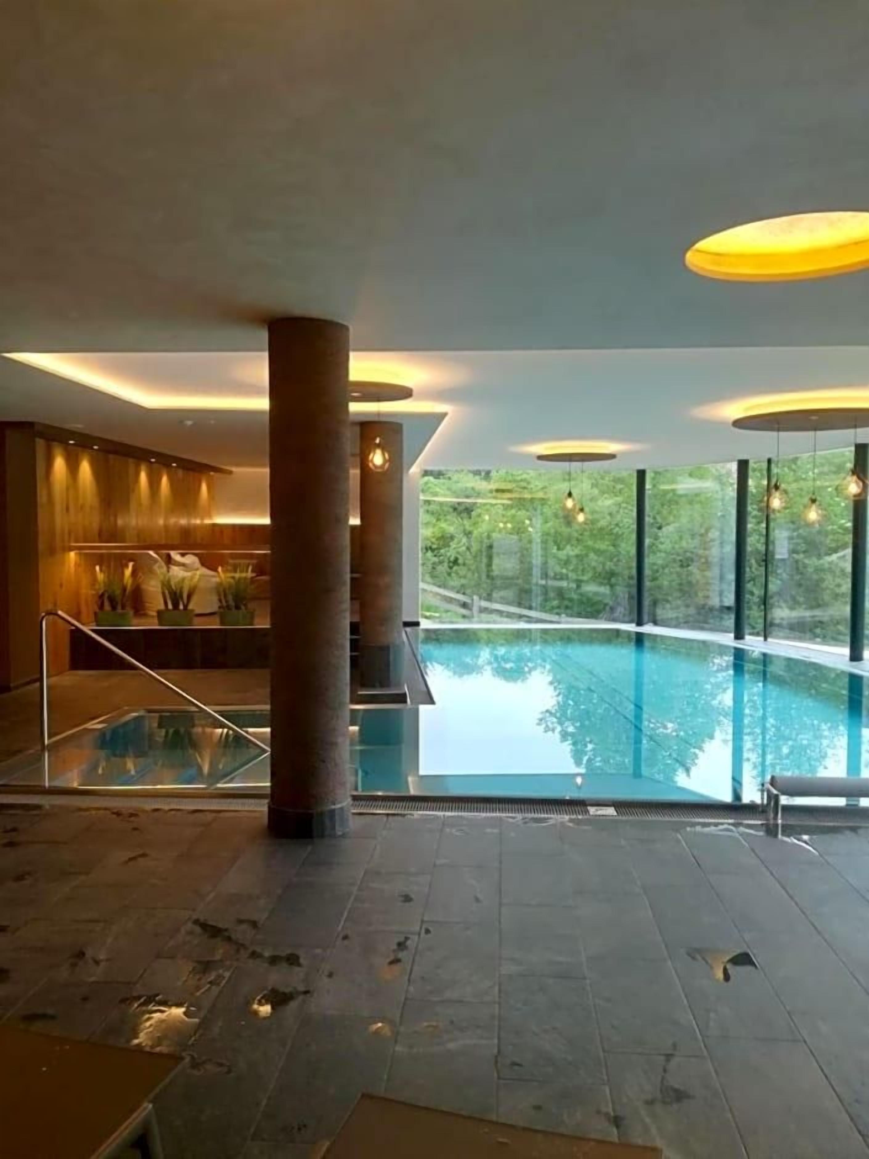 A modern indoor swimming pool with a glass floor, surrounded by large windows and a reception area with plants.