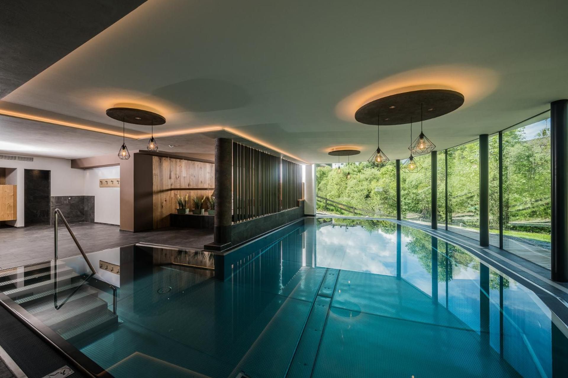 Une piscine intérieure moderne, dotée d’une paroi en verre, de lambris en bois et offrant une vue sur la verdure environnante.