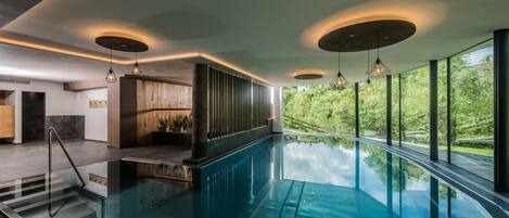 Piscina interior moderna con barandilla de vidrio, paneles de madera y vistas a la vegetación exterior.