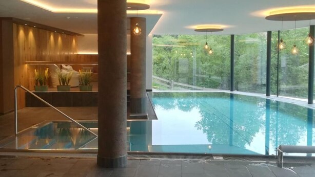 A modern indoor swimming pool with a view of greenery outside, a seating area with a railing, and a wooden wall with a bench.