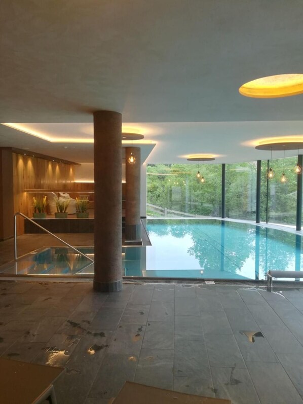 A modern indoor swimming pool with a view of greenery outside, a seating area with a railing, and a wooden wall with a bench.
