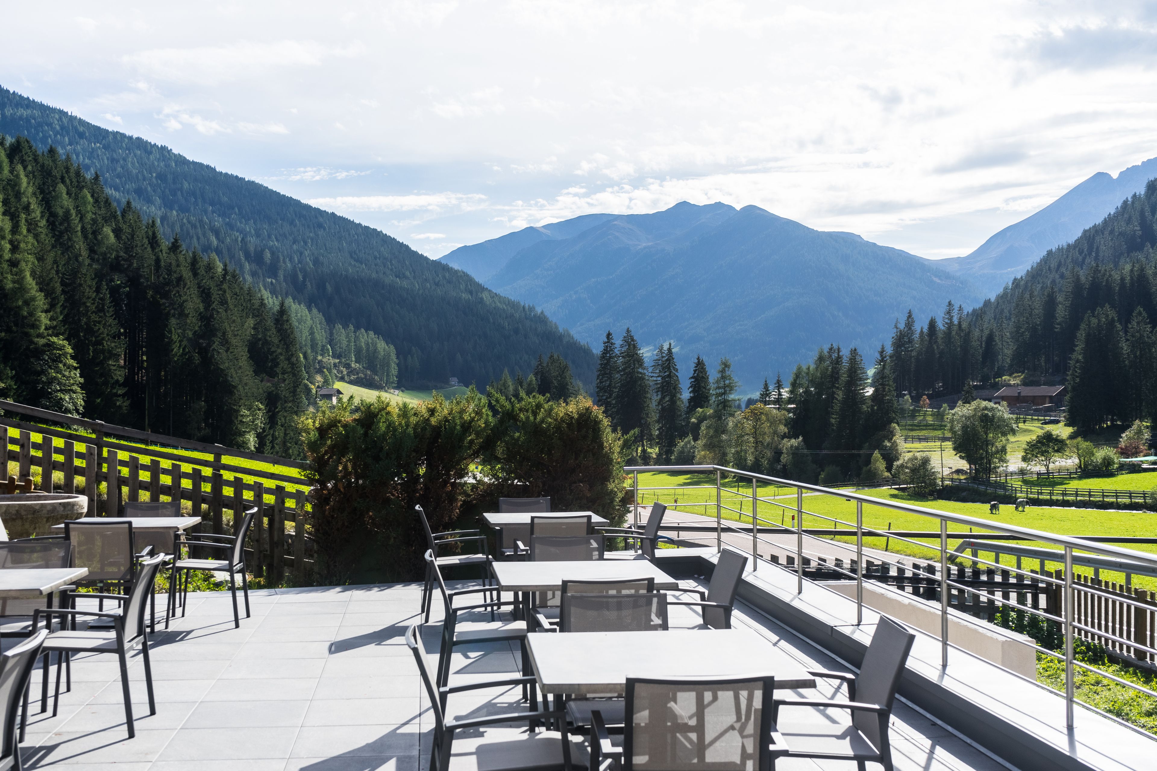 An outdoor terrace with tables and chairs, overlooking a mountainous landscape with a green field and a fence.