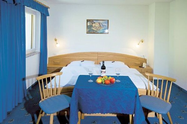 A hotel room with a wooden bed, two blue chairs, a table with fruit and wine glasses, and a painting on the wall. - Hotel & Restaurant Feldrand (Sarentino)