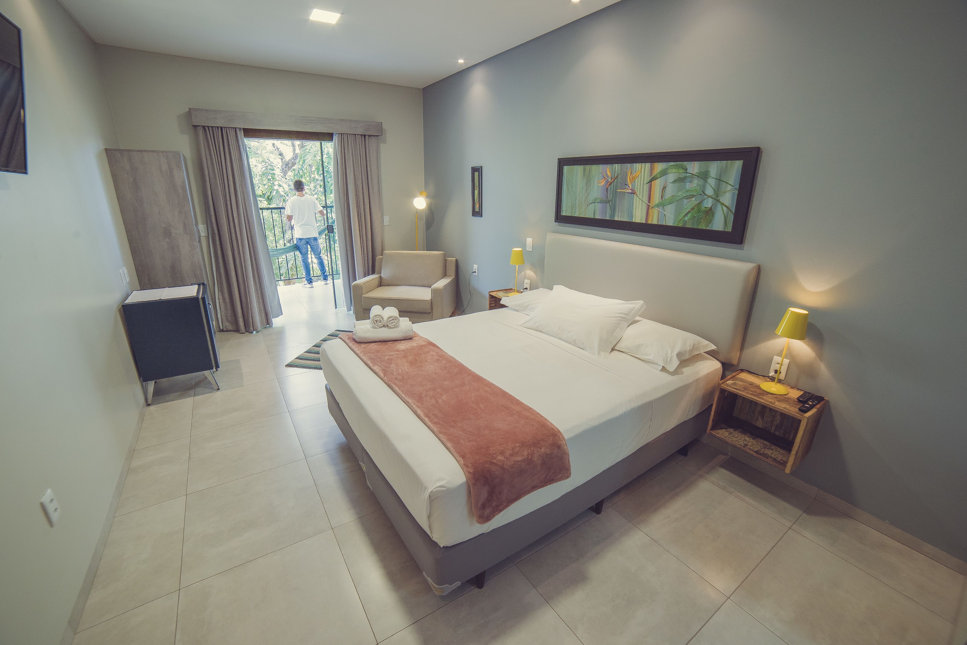 A modern hotel room with a large bed, a chair, a bedside table with a lamp, and a view of the outdoors.