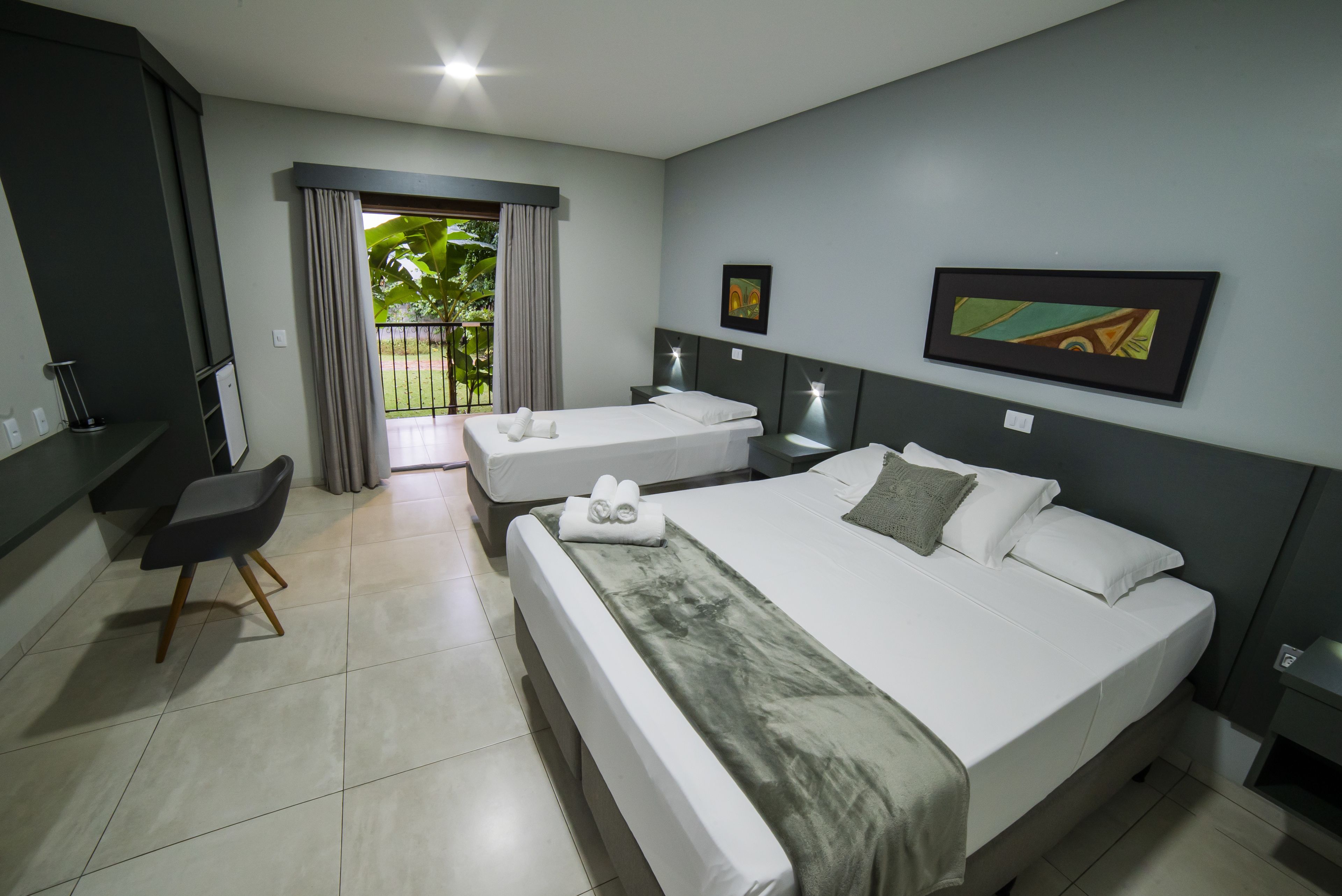 Quarto de hotel com uma cama grande, uma cama menor, uma mesa e uma cadeira. Tem uma varanda com cortinas.