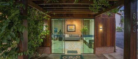 A wooden pergola entrance with a glass door leading to a living area with a couch and a framed picture on the wall.