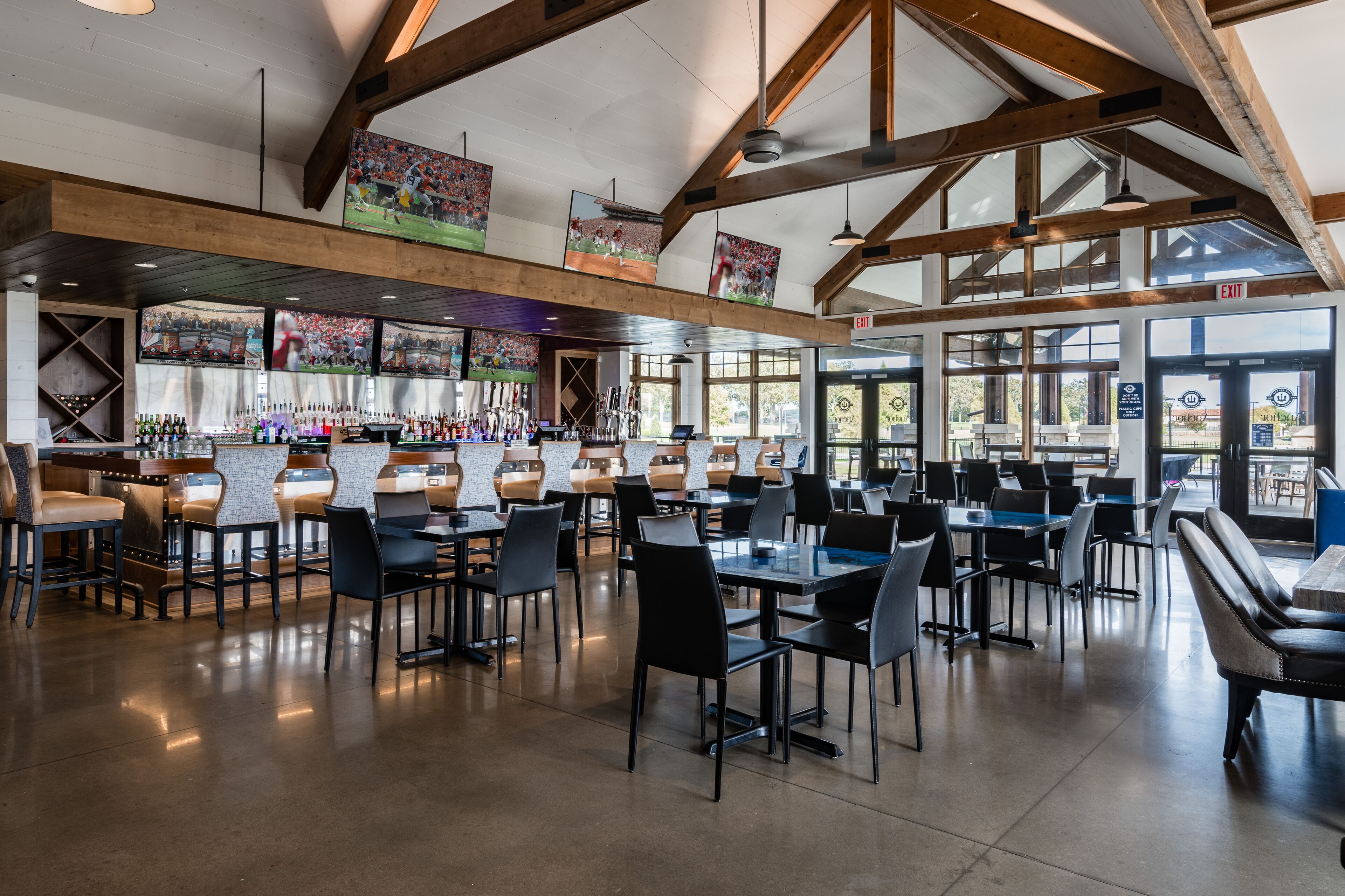 A spacious indoor dining area with high ceilings and exposed beams, featuring multiple tables and chairs, a bar area with a variety of bottles, and large windows allowing natural light.