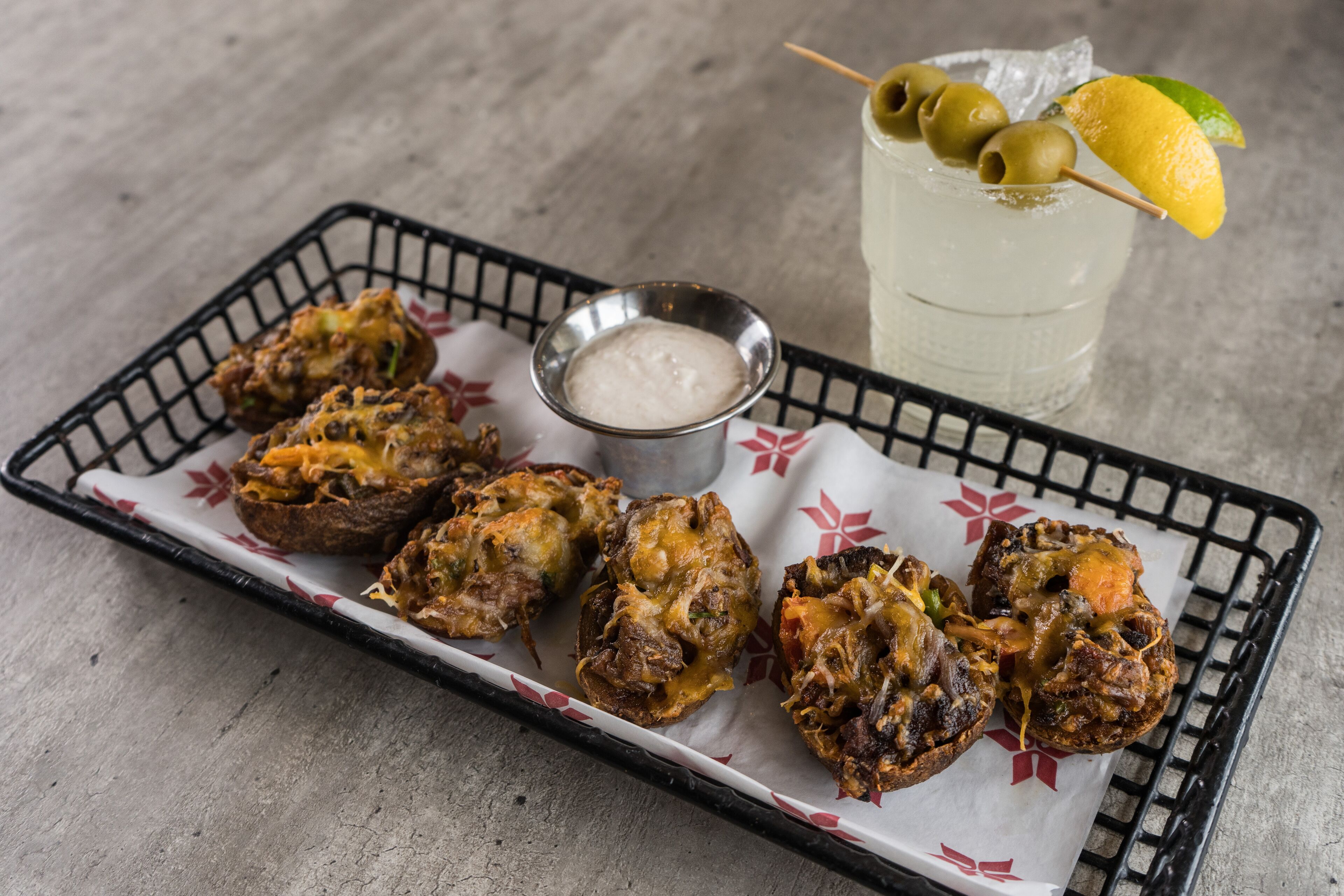 A tray with baked potatoes topped with melted cheese, a small bowl of dipping sauce, and a glass of iced drink with olives and a lemon slice.