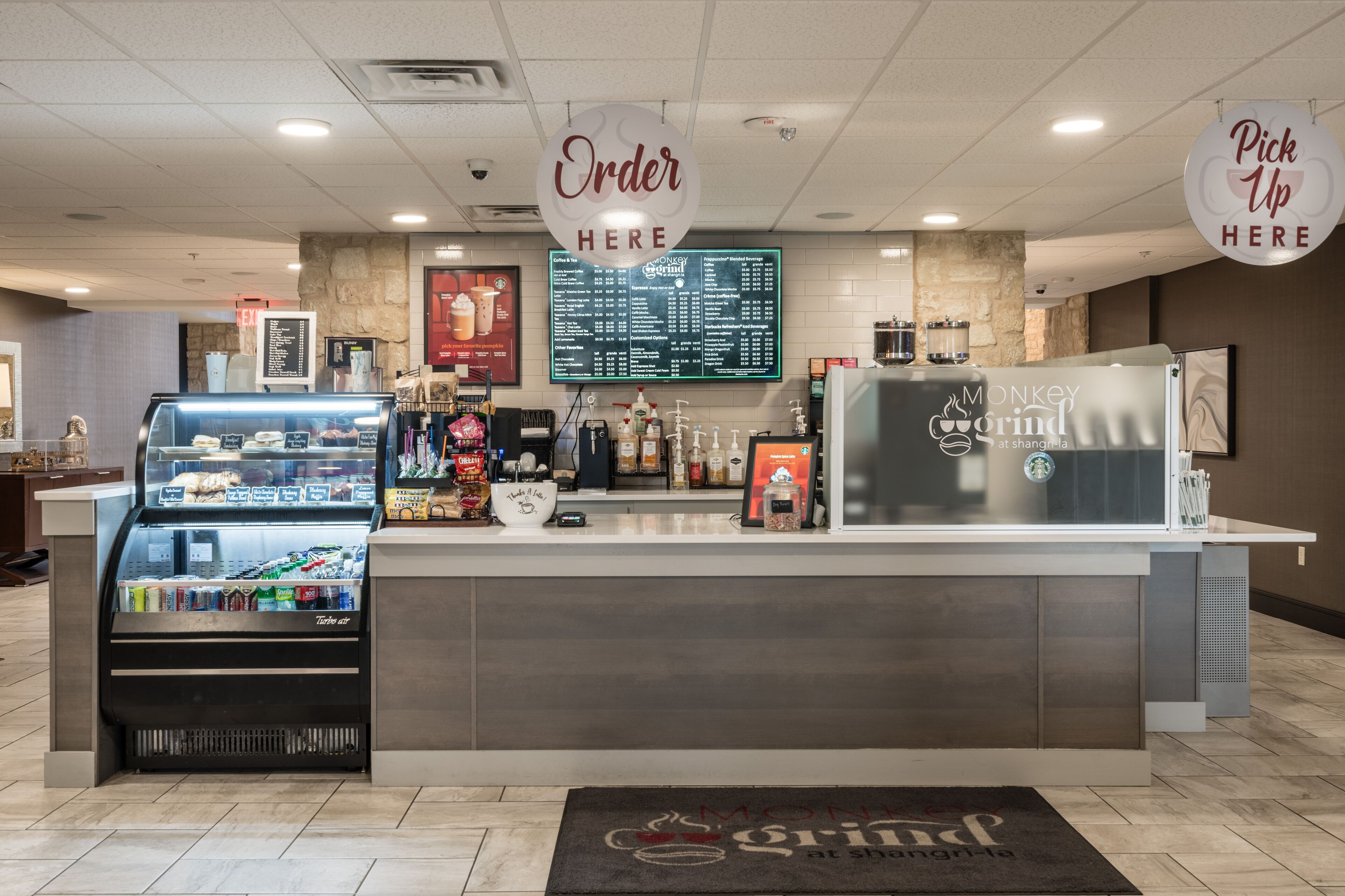 Mostrador de una cafetería con una pizarra de menú y una vitrina refrigerada, y un letrero que indica 'Recoger Aquí'.