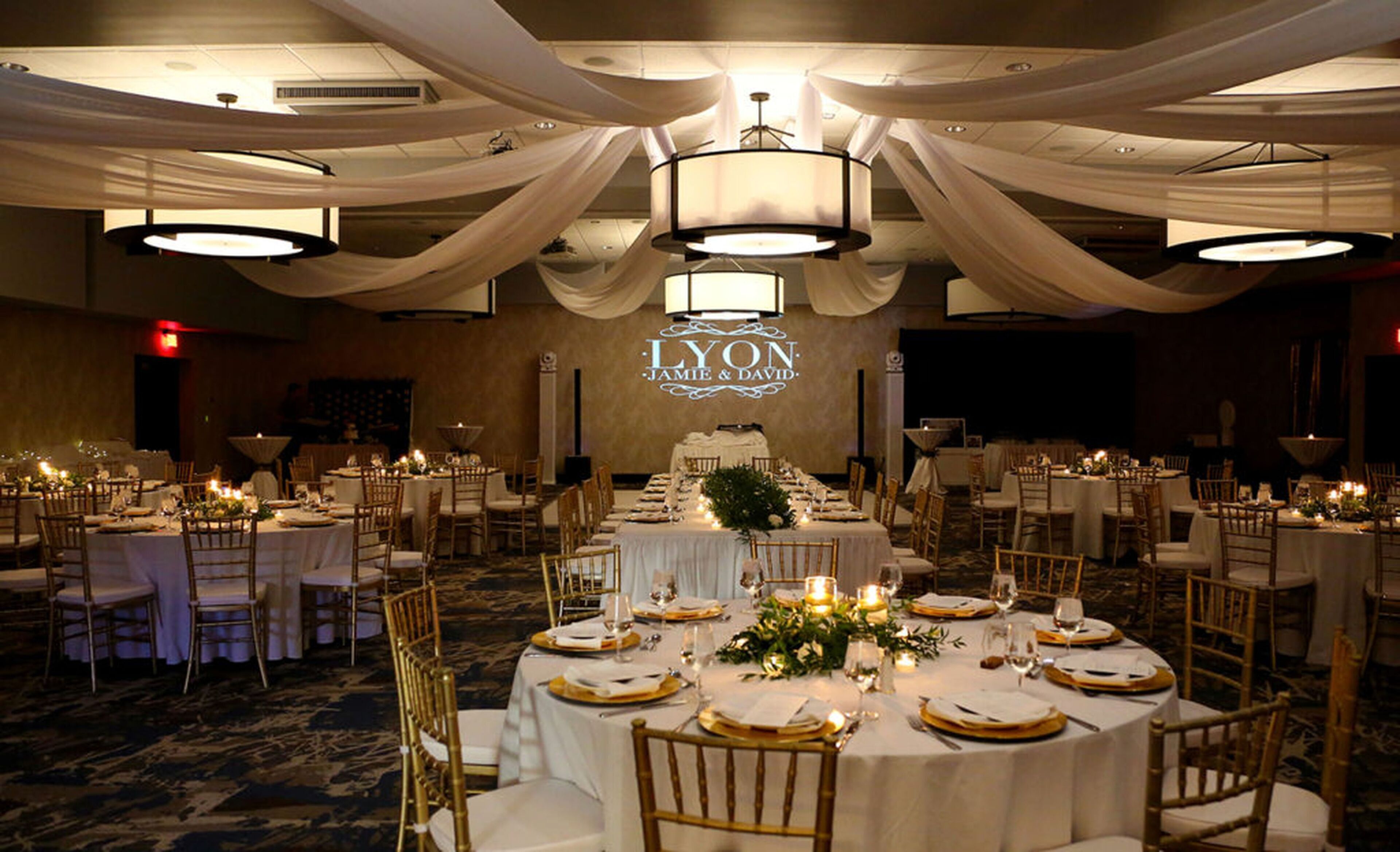 Une salle de banquet avec des tables rondes dressées pour un événement formel, agrémentée de nappes blanches, de touches dorées et d’un éclairage élégant.