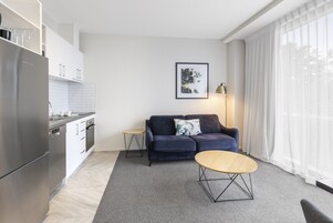 Una cocina moderna con un refrigerador de acero inoxidable, armarios blancos y una pequeña isla. Un sofá azul oscuro con un cojín está ubicado cerca de una ventana con cortinas traslúcidas. Una mesa de centro redonda de madera se encuentra en primer plano.