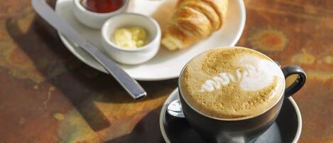 Eine Tasse Kaffee mit Latte-Art auf einem Untertasse, ein Croissant und kleine Schälchen mit Butter und Marmelade auf einem Teller.