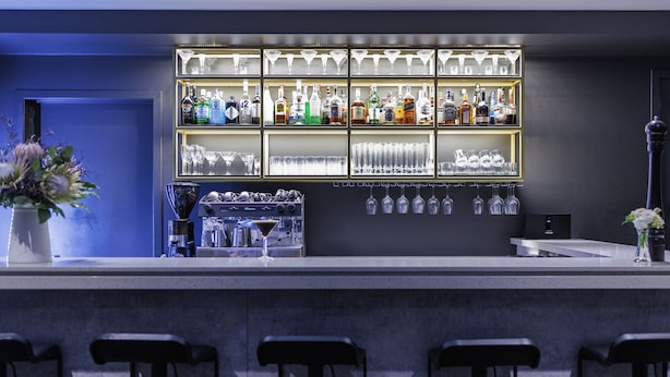 A bar area with a shelf of liquor bottles and glasses, a coffee machine, and bar stools.