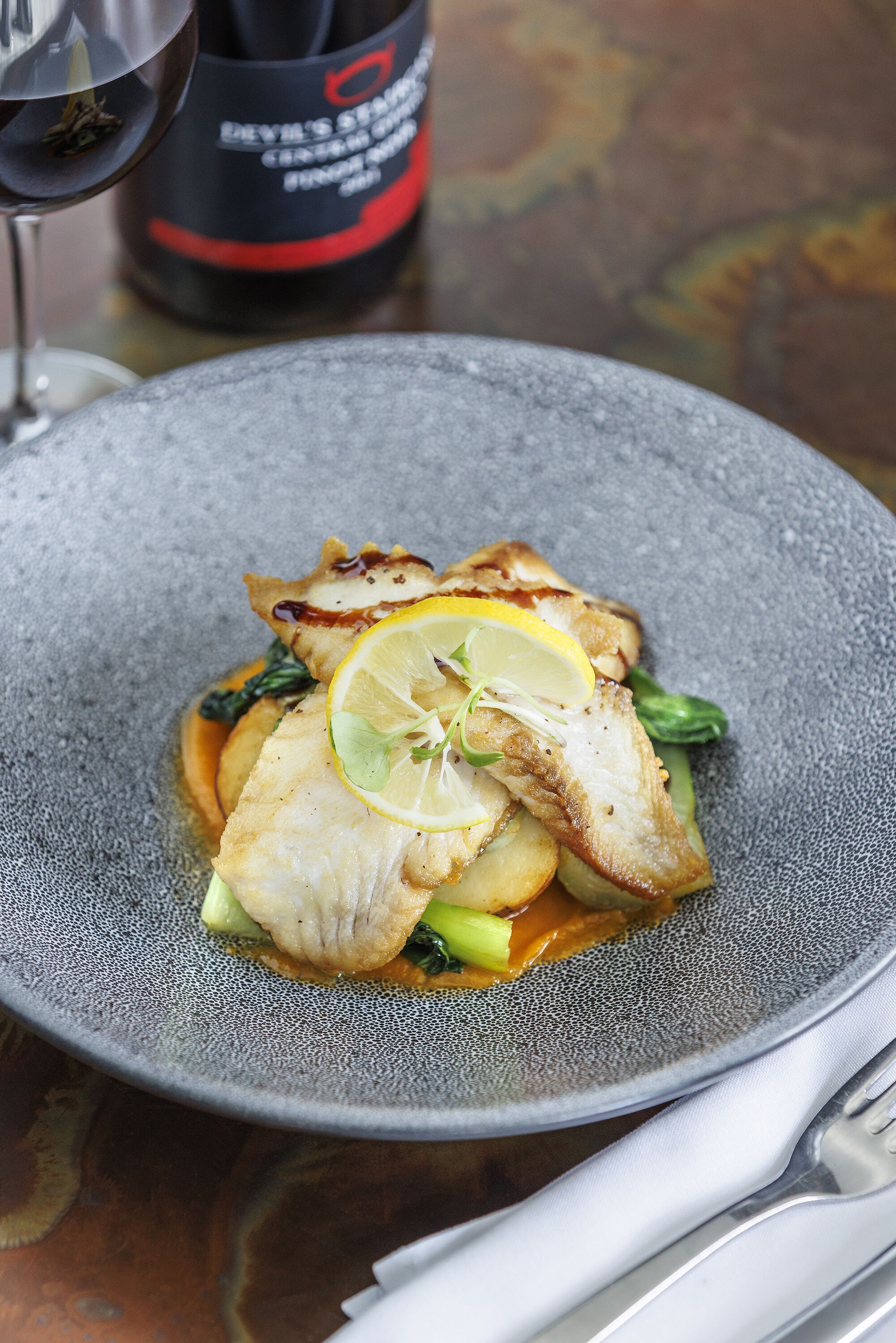 A plate of food with a lemon slice on top, a bottle of wine, and a glass of wine in the background.