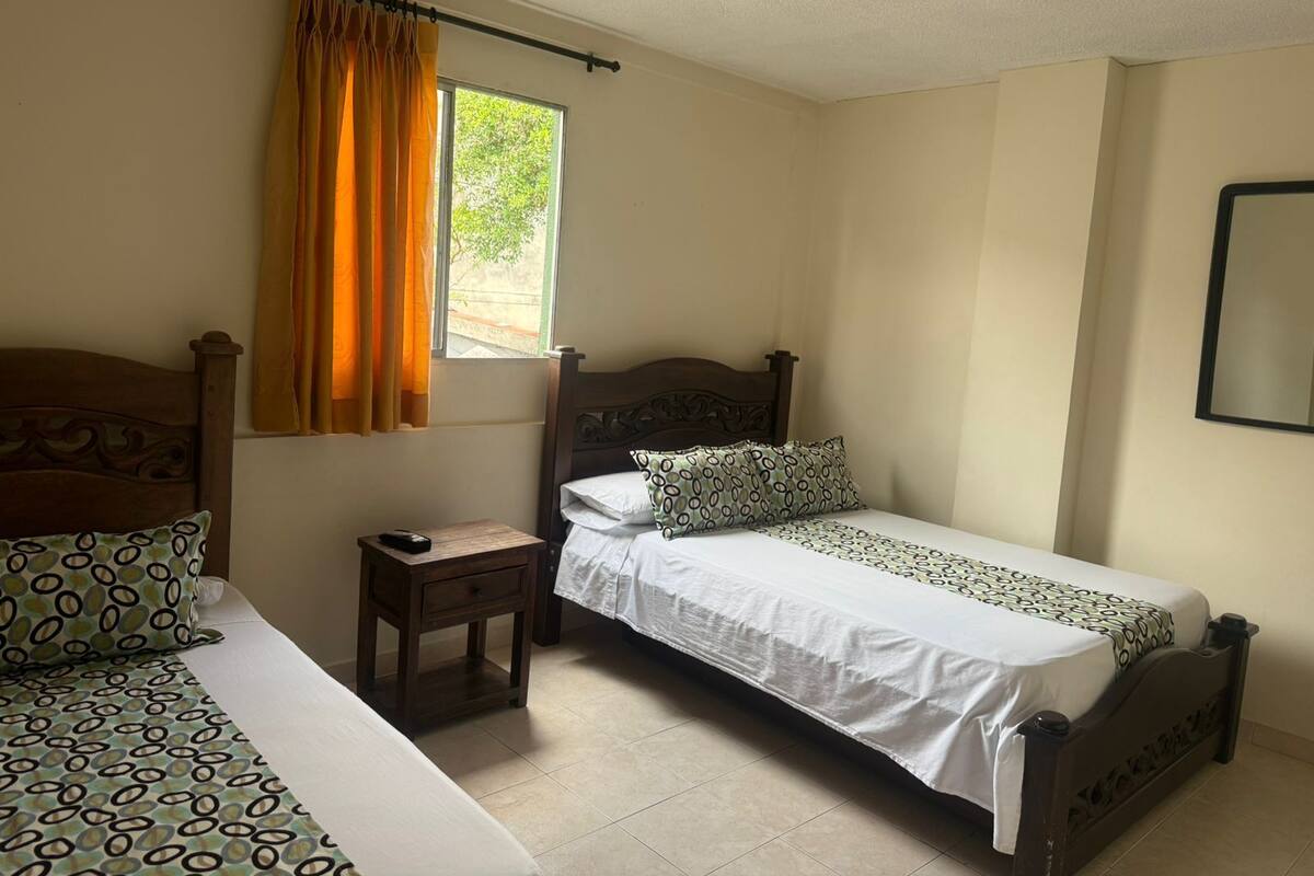 A bedroom with two beds, a nightstand, a window with curtains, and a mirror on the wall.