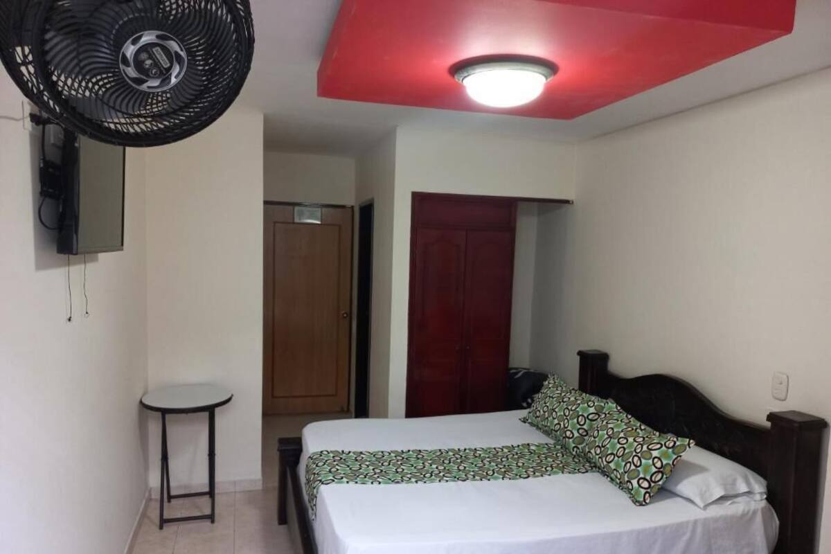 A bedroom with a bed, a standing fan, a small table, and a red ceiling light.