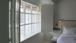 Un dormitorio con un ventanal grande, una cama con ropa de cama blanca, una mesita de noche y un reloj en la pared.