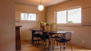 A dining area with a wooden table and four chairs, a hanging light fixture, and windows with curtains.