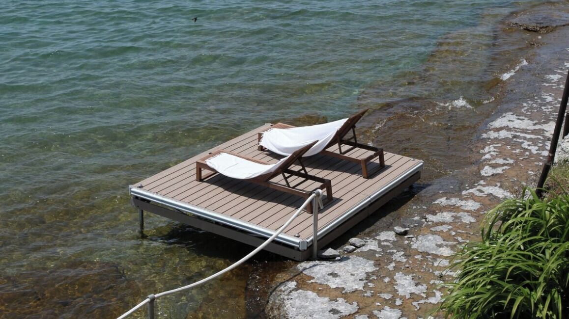 A wooden dock with two lounge chairs and a white cover, extending into a clear body of water.