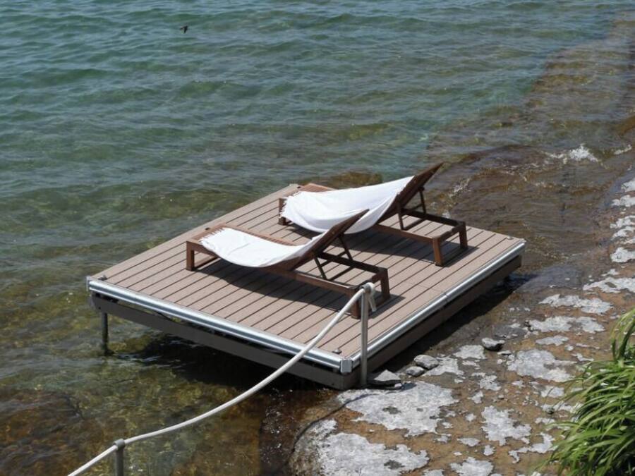 A wooden dock with two lounge chairs and a white cover, extending into a clear body of water.