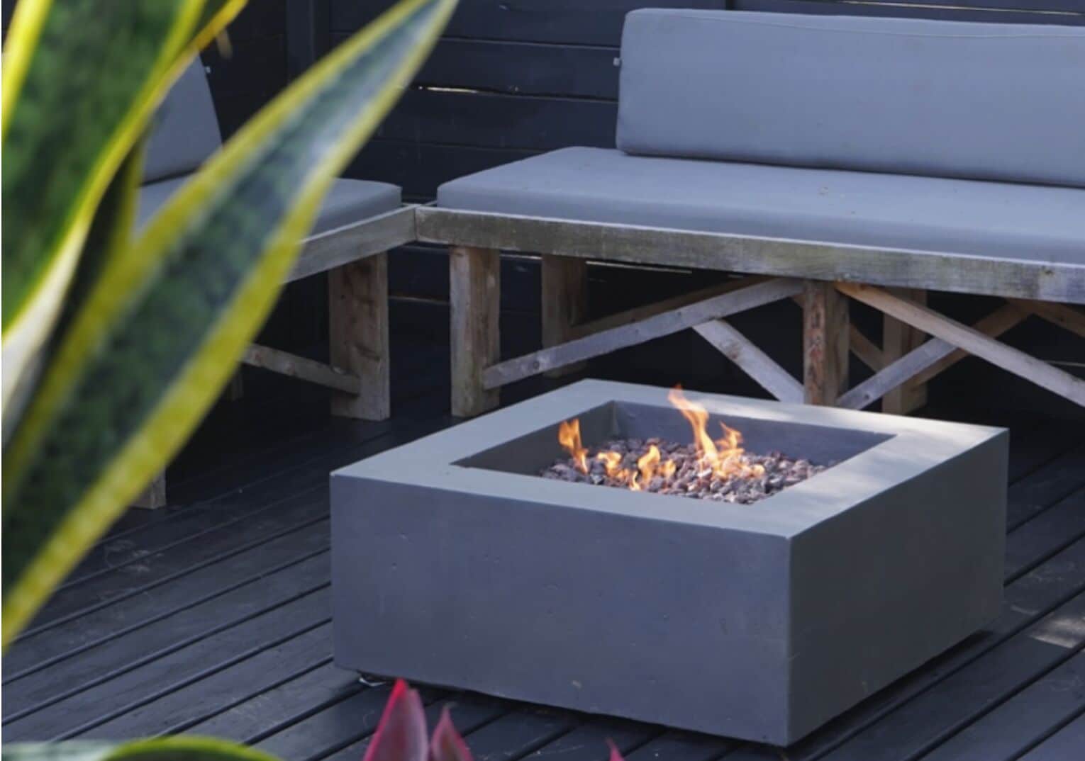A square fire pit with flames, surrounded by wooden benches and a dark wooden deck.