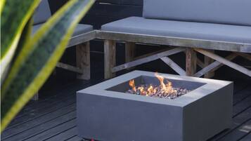 A square fire pit with flames, surrounded by wooden benches and a dark wooden deck.