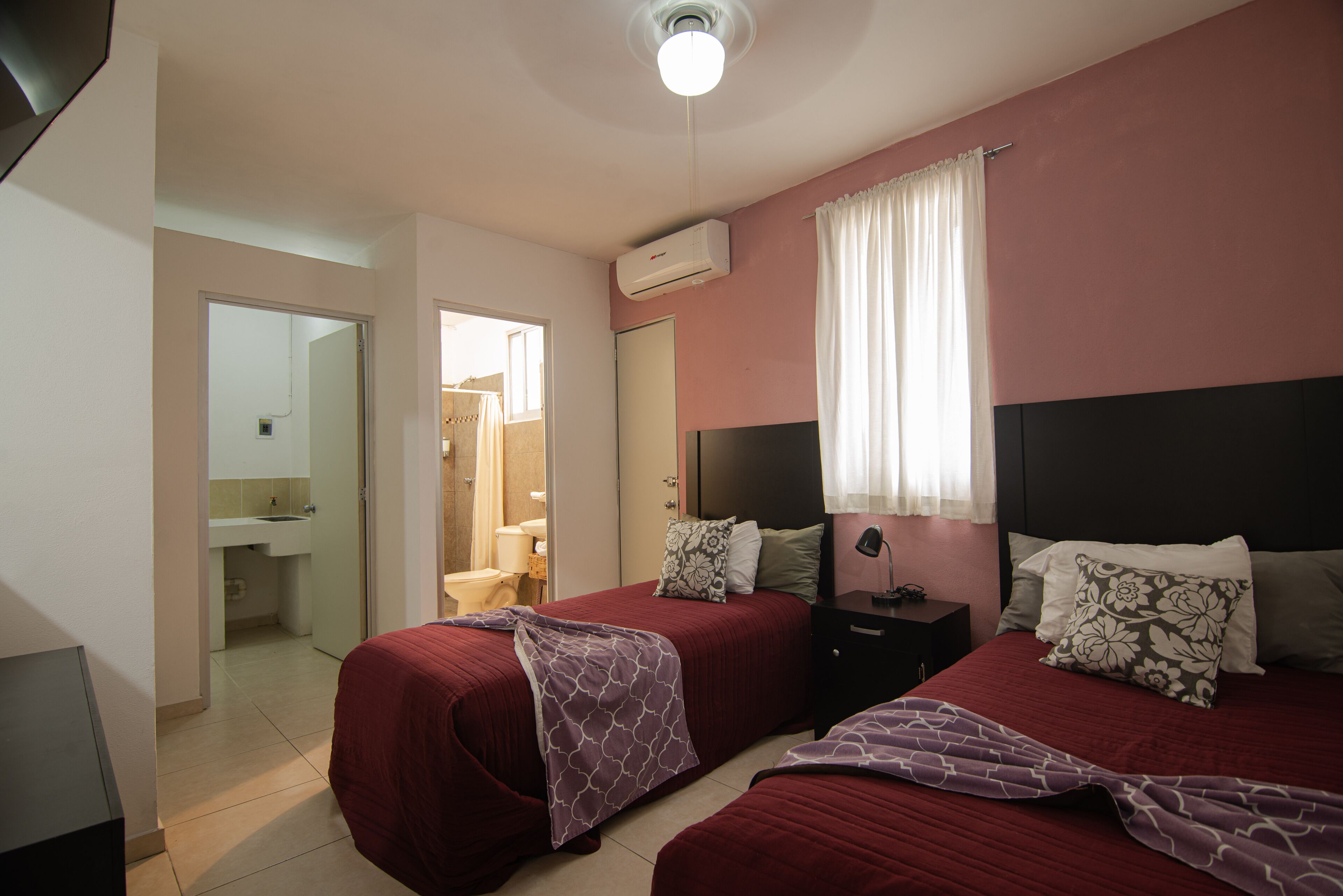 Un dormitorio con dos camas, una mesita de noche, un escritorio, una silla y un baño visible a través de una puerta abierta.