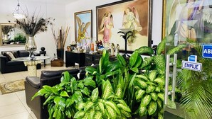 A living room with a sofa, coffee table, and potted plants. There are paintings on the wall and a ceiling fan.
