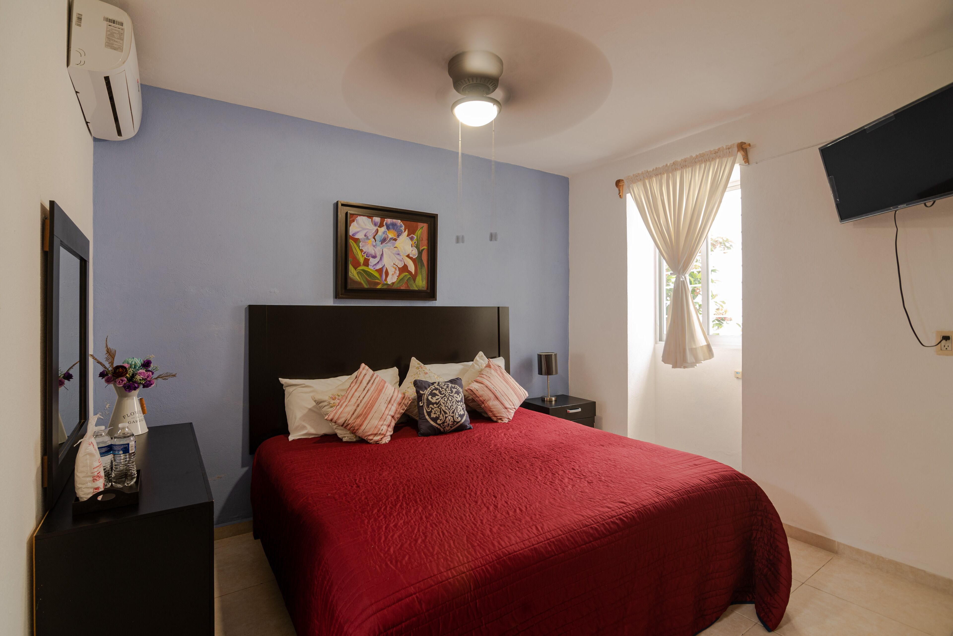 Un dormitorio con una colcha roja, un ventilador de techo, un televisor montado, una mesita de noche con lámpara y una ventana con cortinas.
