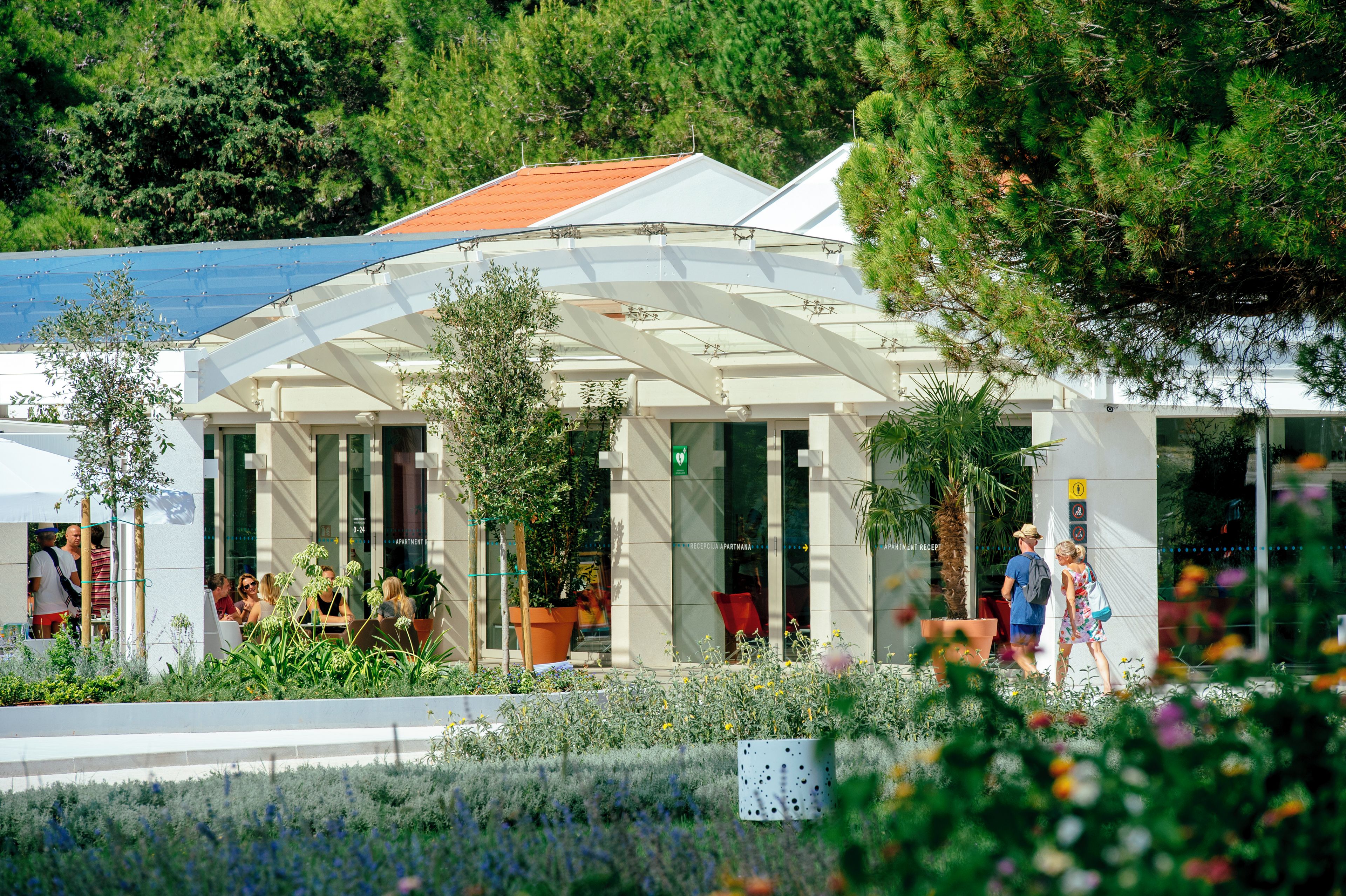 A modern building with a covered entrance, potted plants, and a sign indicating a reception area.