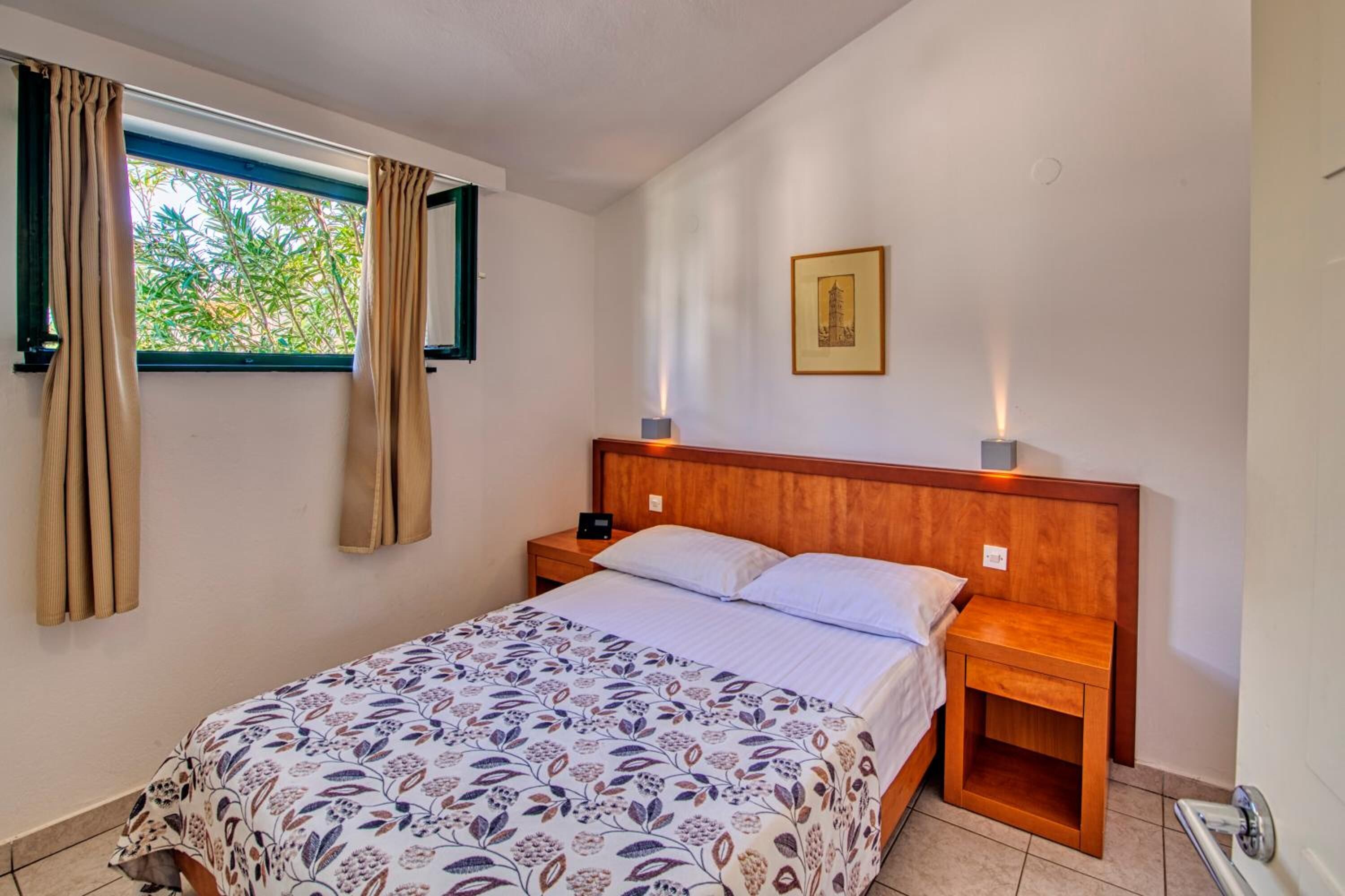 Une chambre à coucher comprenant un lit, des tables de chevet, une fenêtre avec des rideaux, un tableau accroché au mur et un sol carrelé.