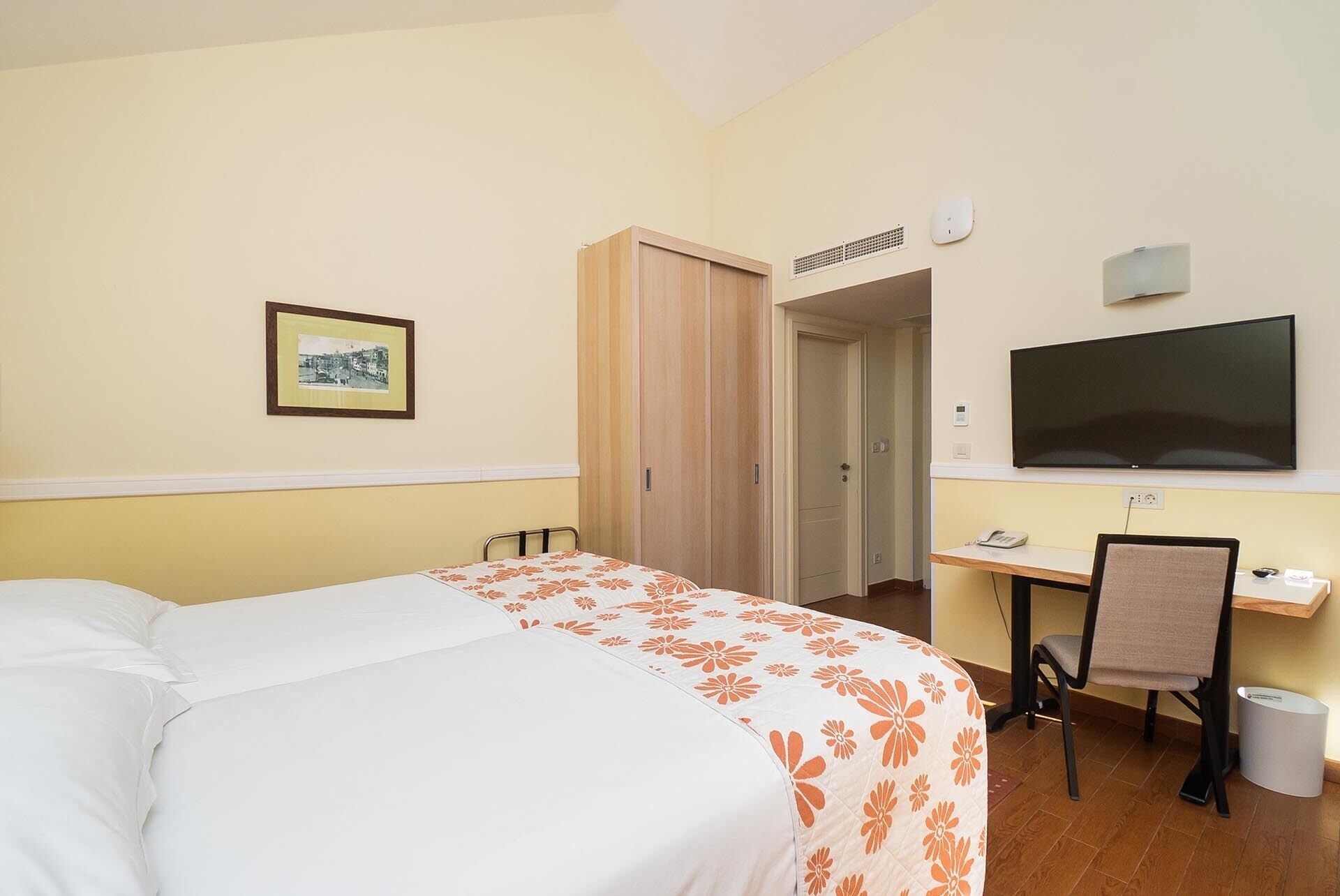 A hotel room with a bed, a desk with a chair, a television, and a framed picture on the wall.