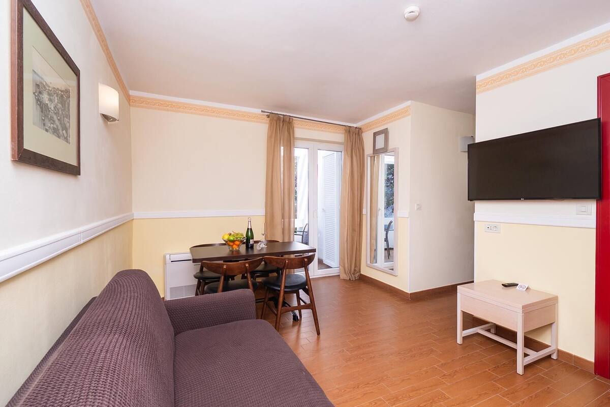 Un salón con sofá, una mesa de comedor con sillas, un televisor montado en la pared y una pequeña mesa con una silla.