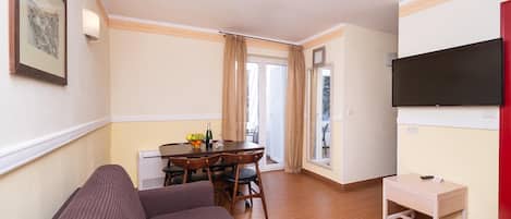 A living room with a sofa, a dining table with chairs, a TV mounted on the wall, and a small table with a chair.