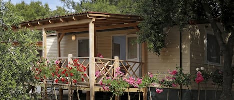 Una cabaña de madera con un porche, rodeada de plantas con flores y árboles.